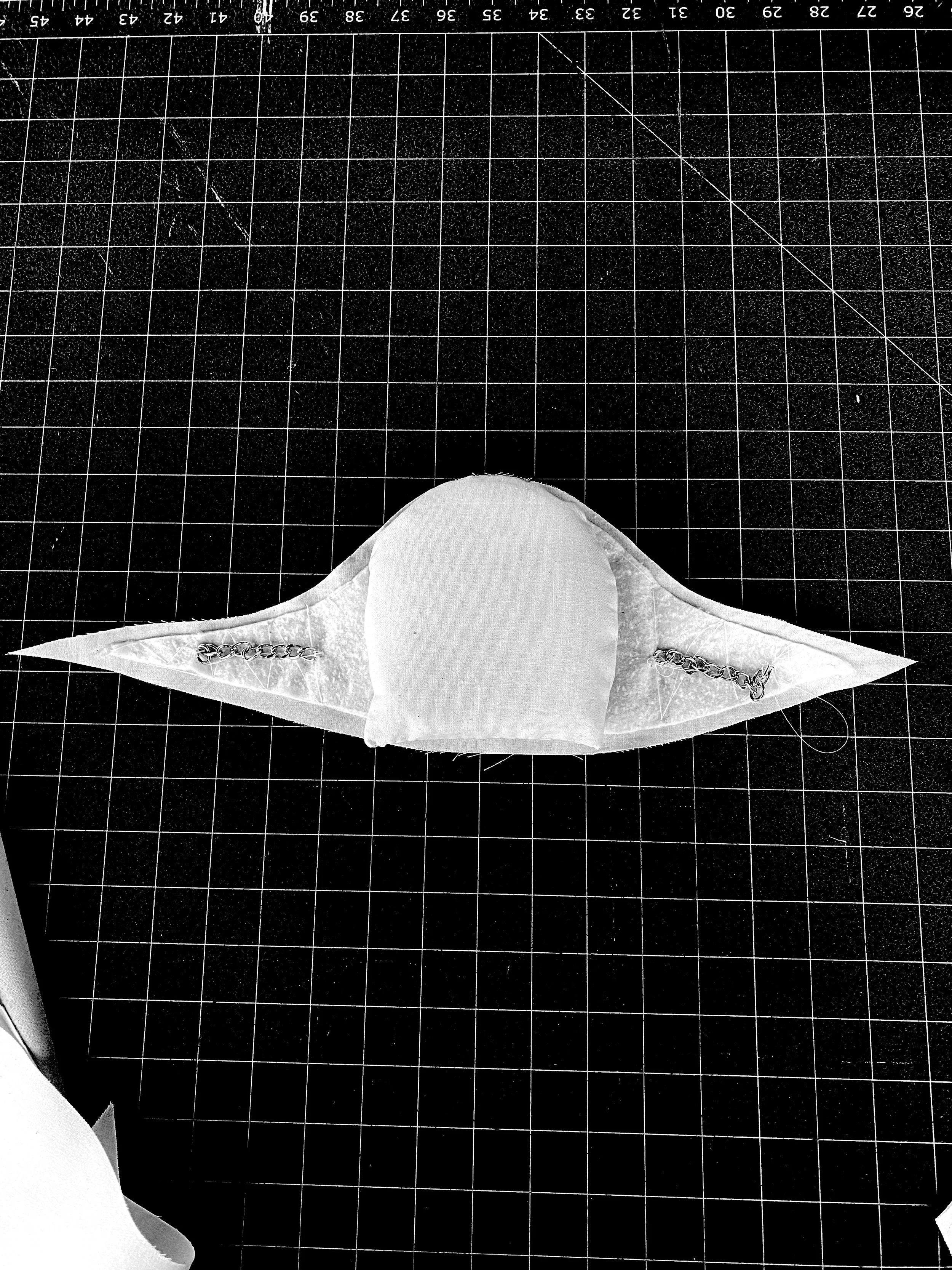 A white fabric cat face with stitched facial features and chains on either side, placed on a black cutting mat with white grid lines.