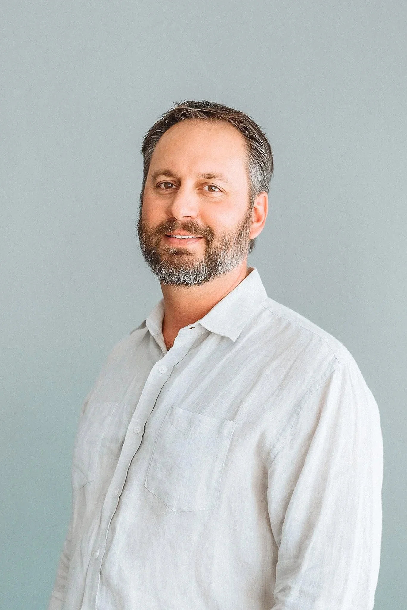 Clay Conrad is wearing a white button-up shirt, smiling slightly and looking at the camera, standing against a plain light gray background.