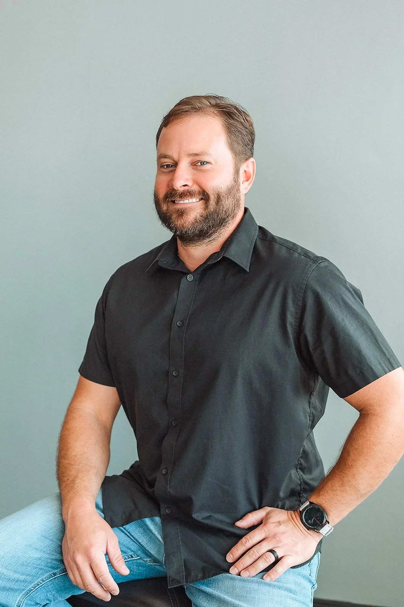 Brent Crowell sitting against a plain light-colored wall, wearing a black button-up shirt, jeans, a wristwatch, and a ring, smiling slightly and looking at the camera.