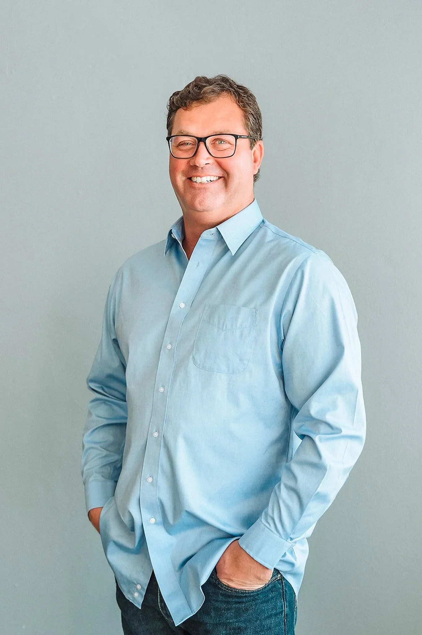 Bain McEldowney, smiling, wearing glasses, a light blue button-up shirt, and jeans, standing against a plain gray wall.