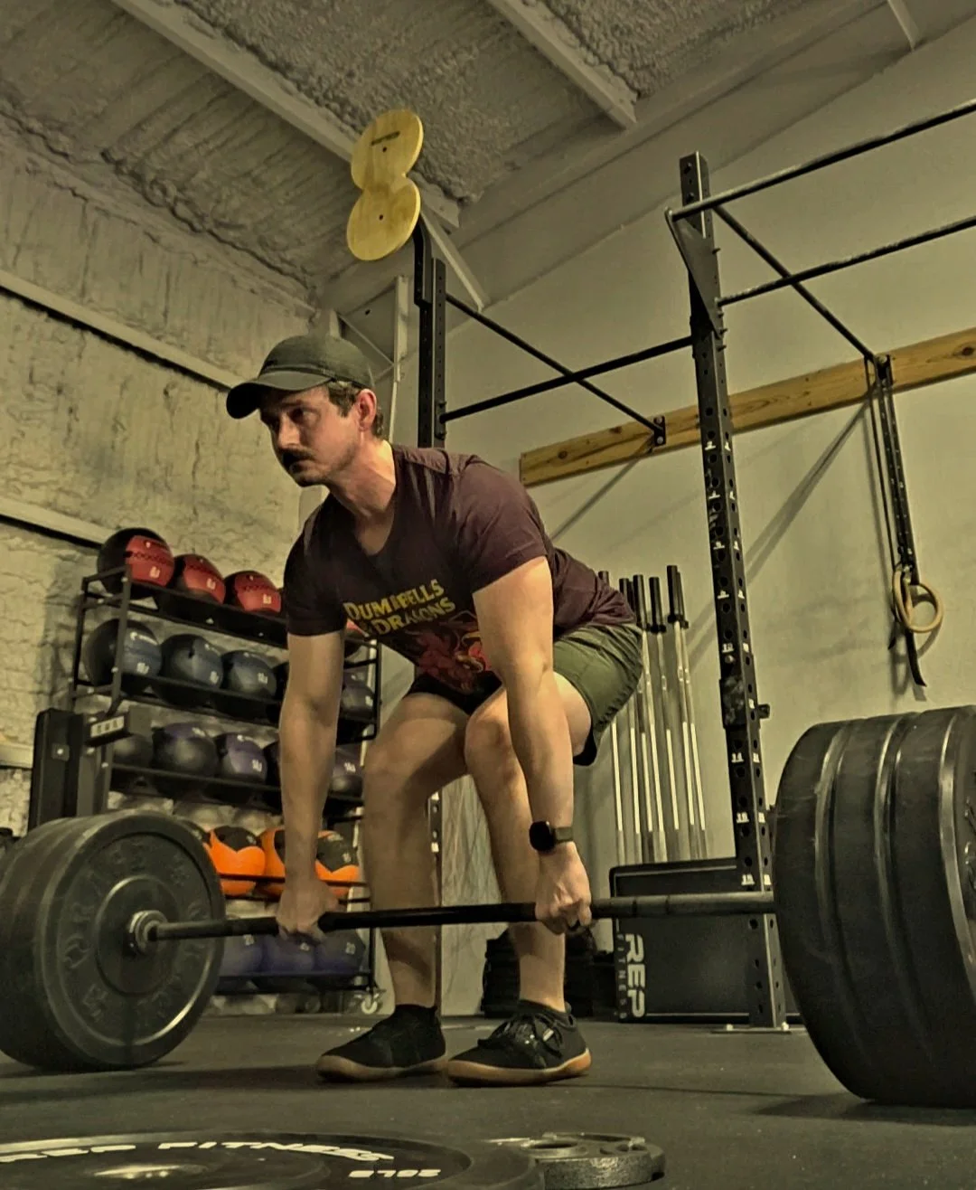A man in a gym preparing to lift a barbell loaded with weights, wearing a dark T-shirt, shorts, a cap, and athletic shoes.