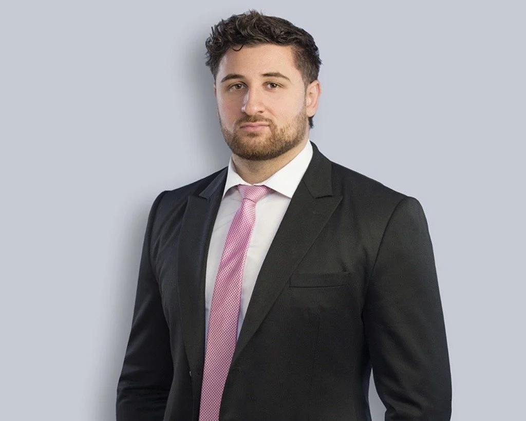 Young man in a black suit with a white shirt and pink tie, standing against a light gray background.