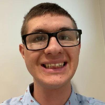 Close-up of a young man with short brown hair, glasses, and a big smile, wearing a light blue shirt with small patterns, standing against a plain background.