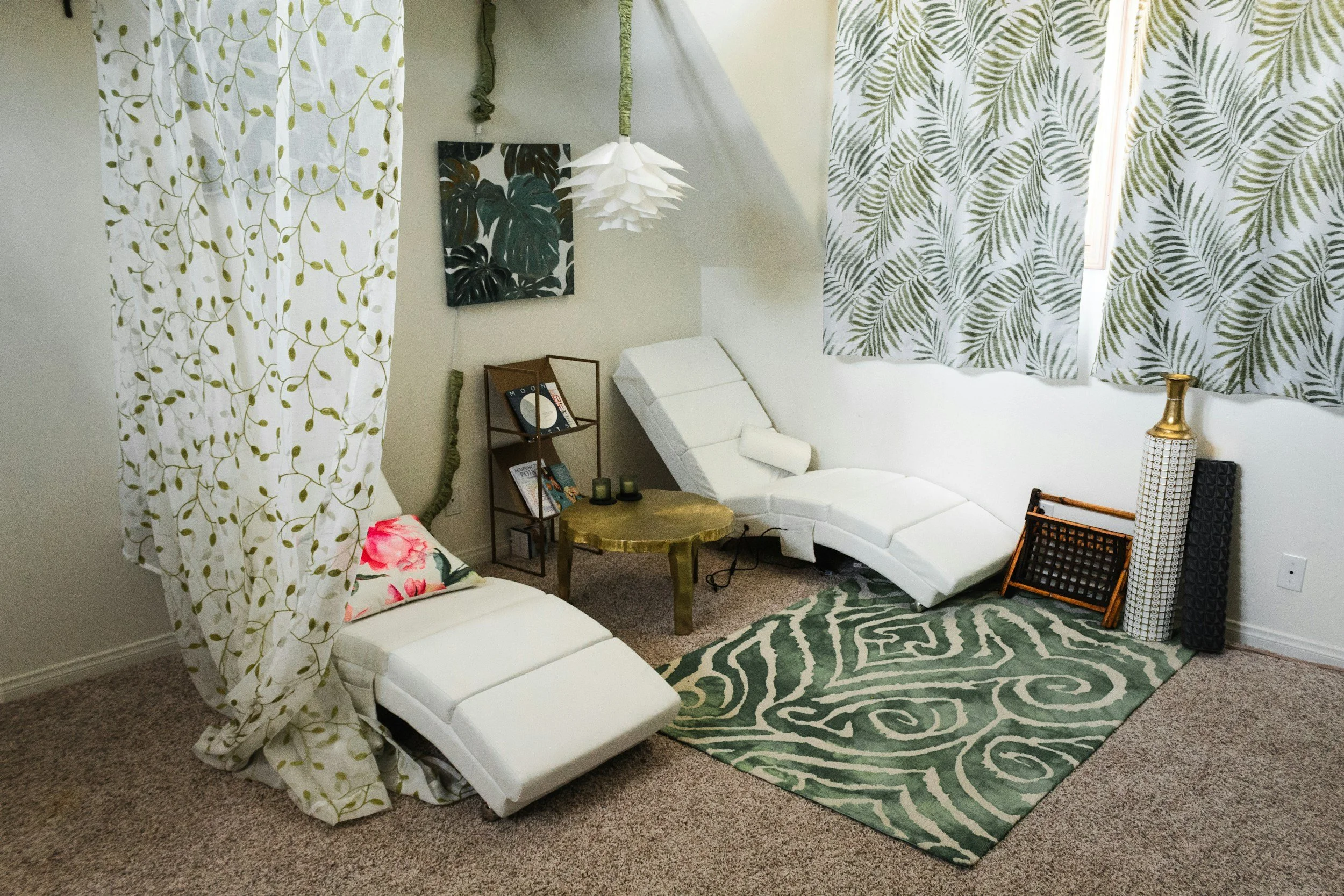 A cozy living room with a green and white color scheme, featuring a white curved lounge chair, a small wooden side table, a patterned area rug, leafy curtains, and decorative wall art with a tropical leaf design.
