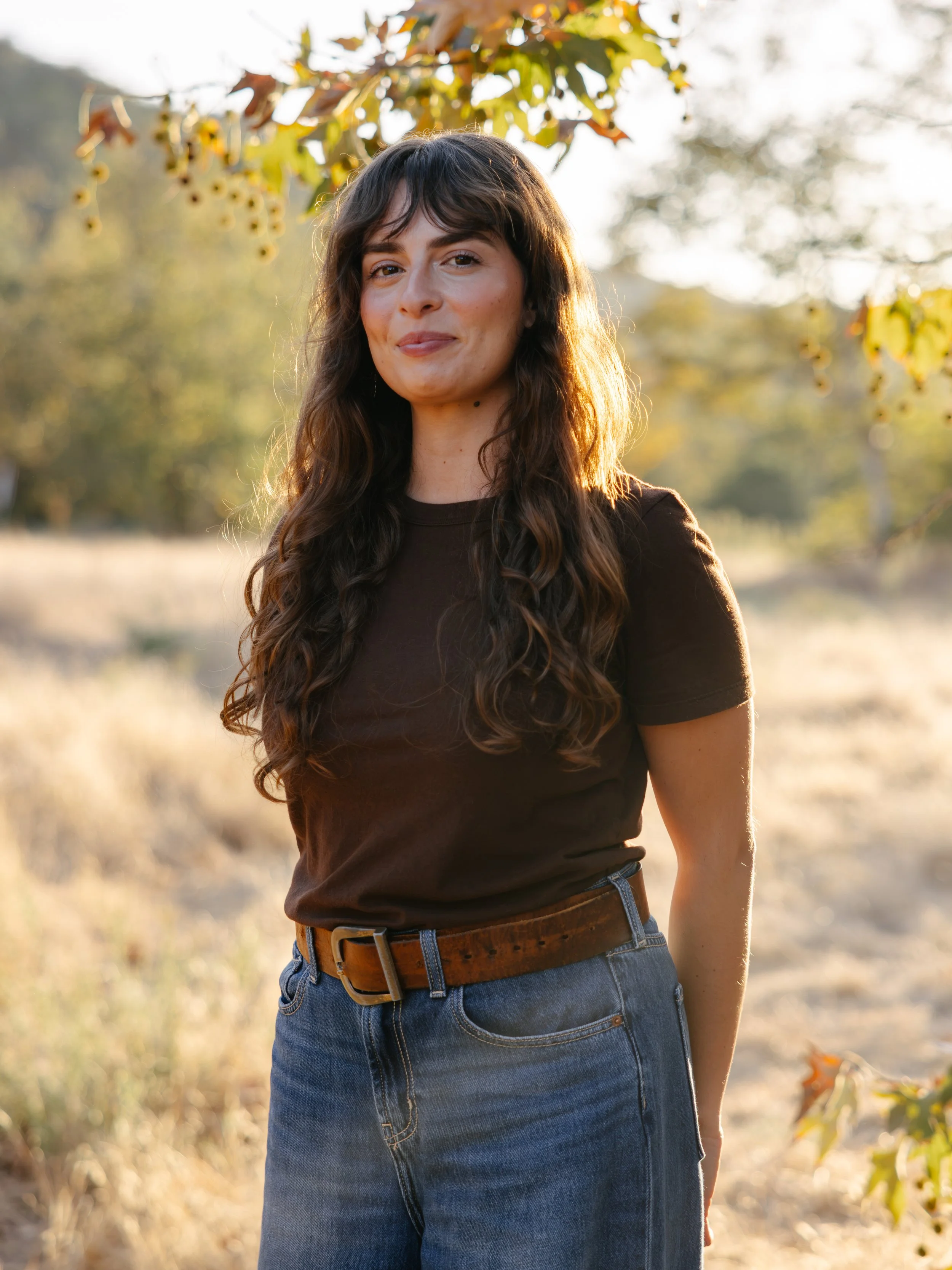 A woman with long curly brown hair wearing a black t-shirt and blue jeans standing outdoors in a field with trees and fallen leaves, illuminated by warm sunlight.