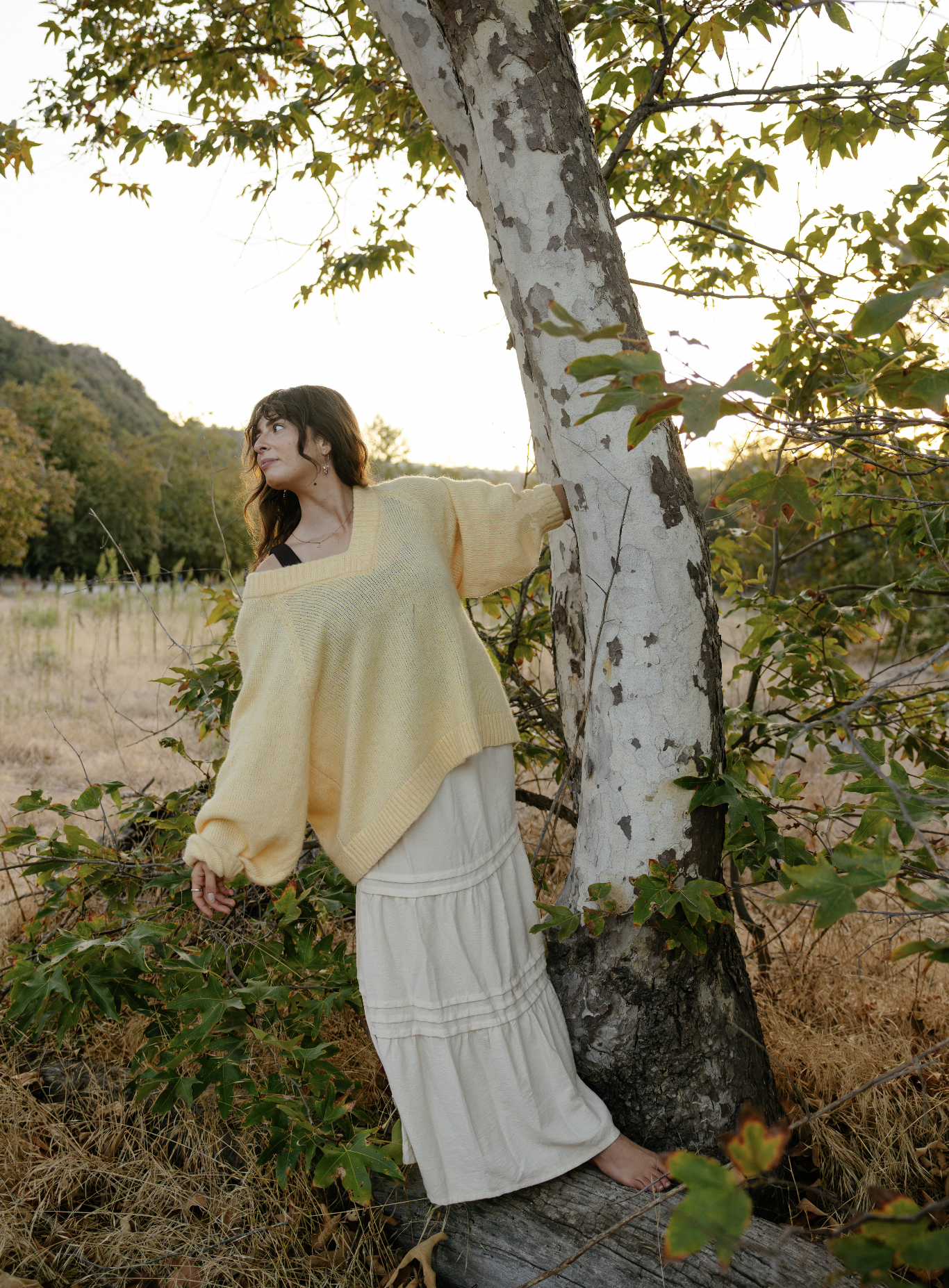 Julianna Stein dark hair leaning against a tree trunk outdoors during daytime, wearing a loose yellow sweater and a long cream-colored skirt, surrounded by greenery and dry grass, with sunlight filtering through the leaves.