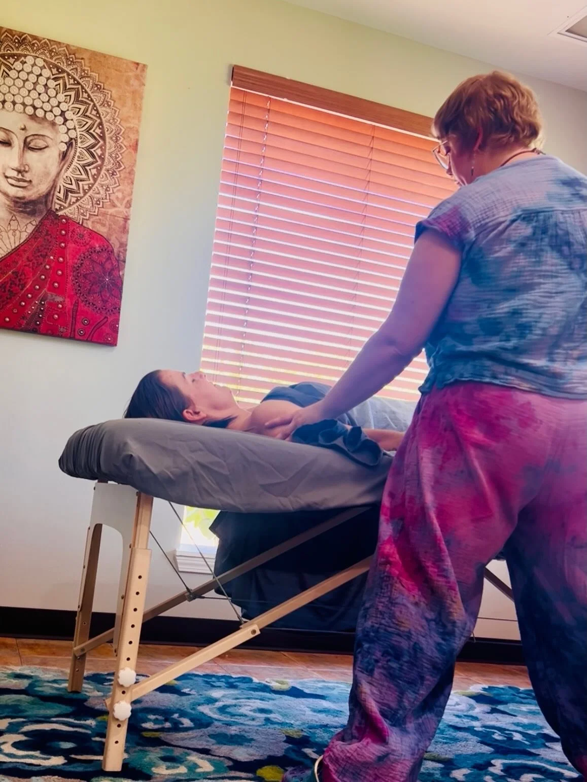 A woman lying on a massage table with her eyes closed, receiving a massage from a therapist. The room has a window with wooden blinds, a colorful patterned rug, and a painting of Buddha on the wall.