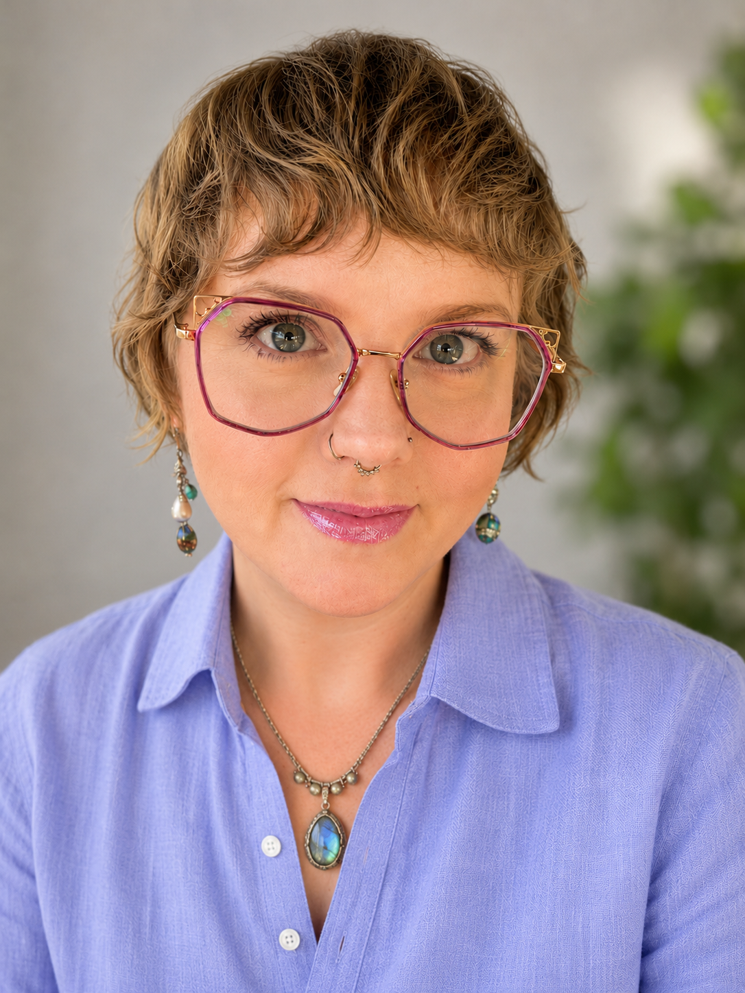 A woman with short, wavy brown hair wearing large pink glasses, a lavender shirt, and jewelry with a large blue stone pendant and dangling earrings, standing in front of a blurred green plant background.