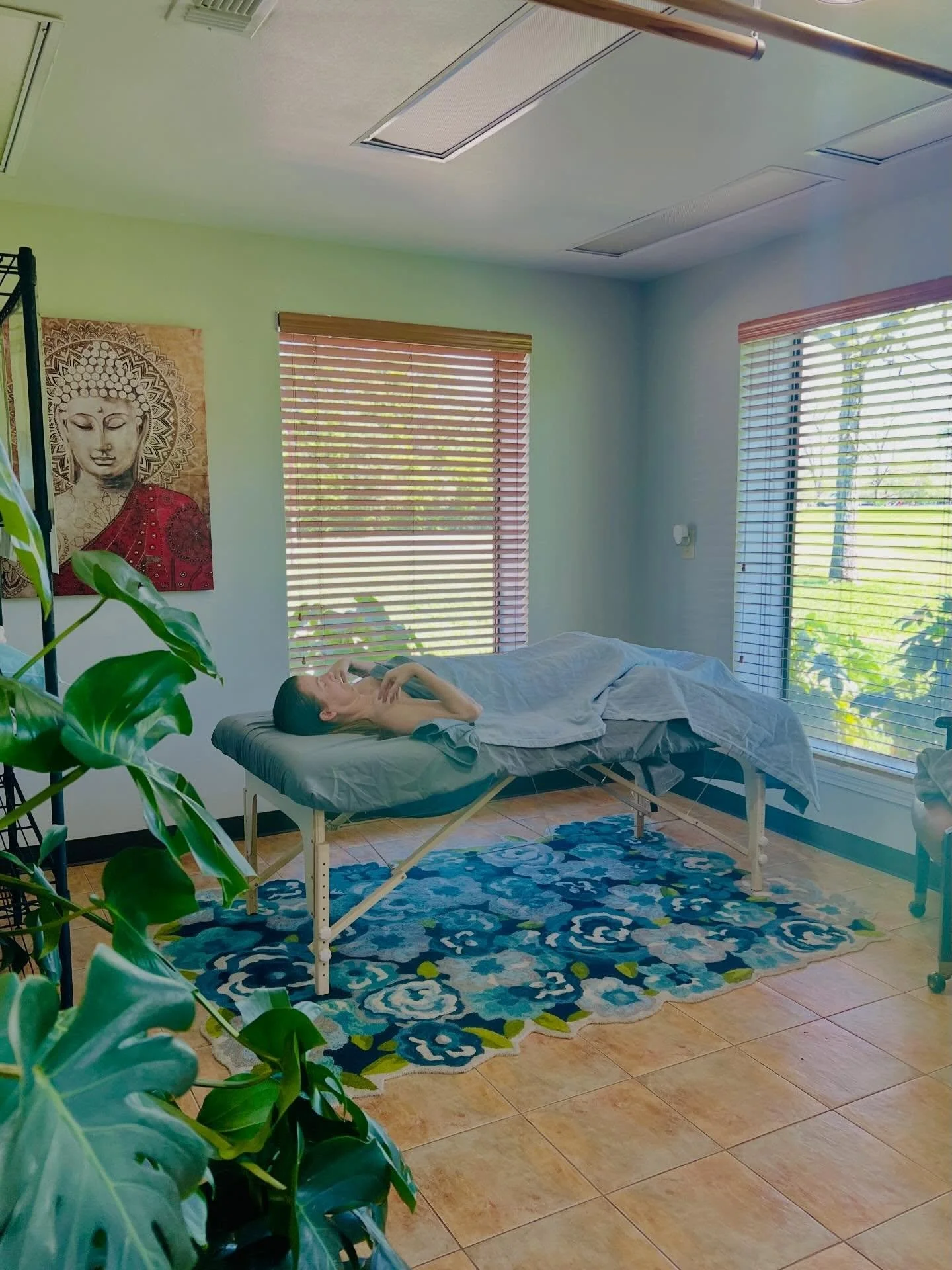 A person lying on a massage table in a bright room with large windows, green walls, a Buddha artwork on the wall, a blue patterned rug, and green plants.