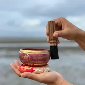 Person holding a wooden percussion mallet above a colorful singing bowl on a person's hand, with a cloudy sky and ocean background.