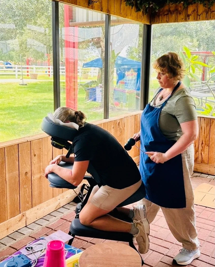 A woman is receiving a massage on a massage chair from a massage therapist while inside a wooden-paneled room with large windows overlooking a grassy outdoor area.