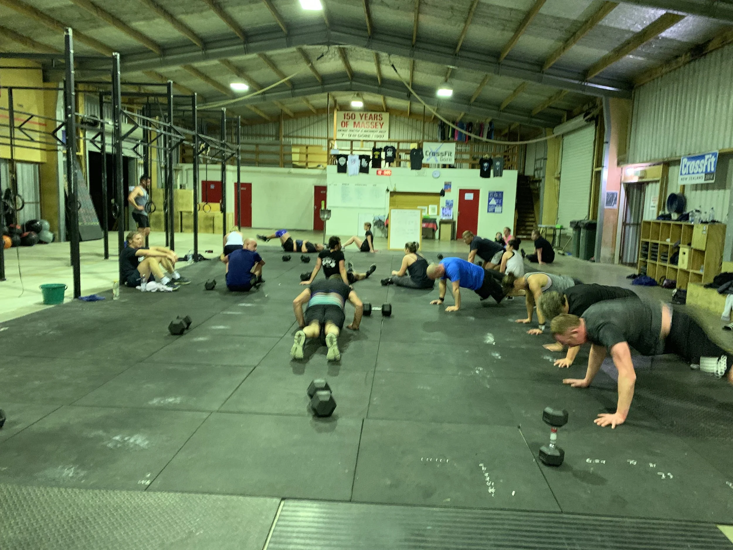 People participating in a group workout or class in a gym, performing exercises on the floor with some standing, surrounded by gym equipment and a sign that reads '150 Years of Massey'.
