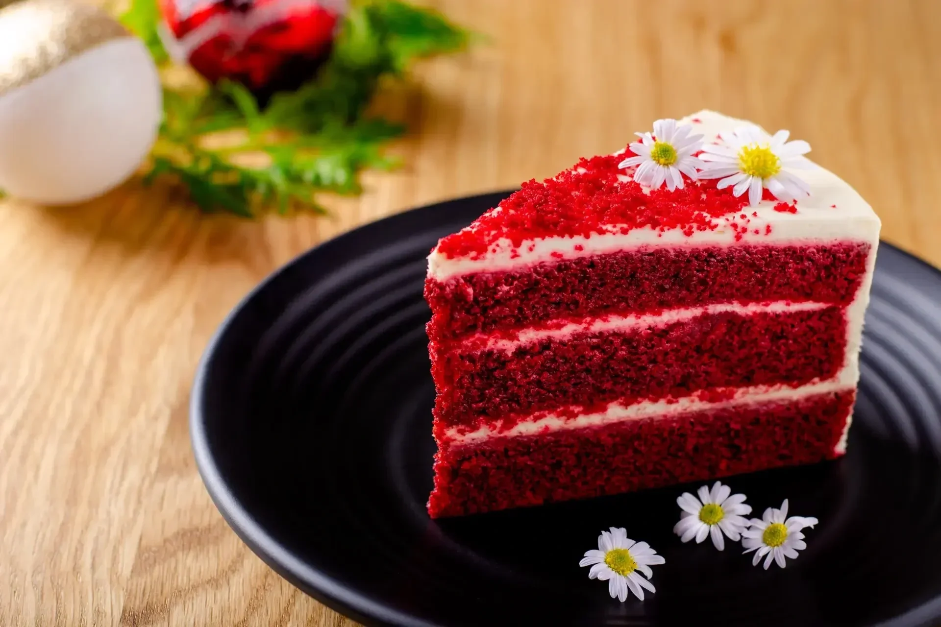 A slice of red velvet cake with white frosting, decorated with small white flowers, on a black plate on a wooden surface, with a blurred background including a white container and red flower.