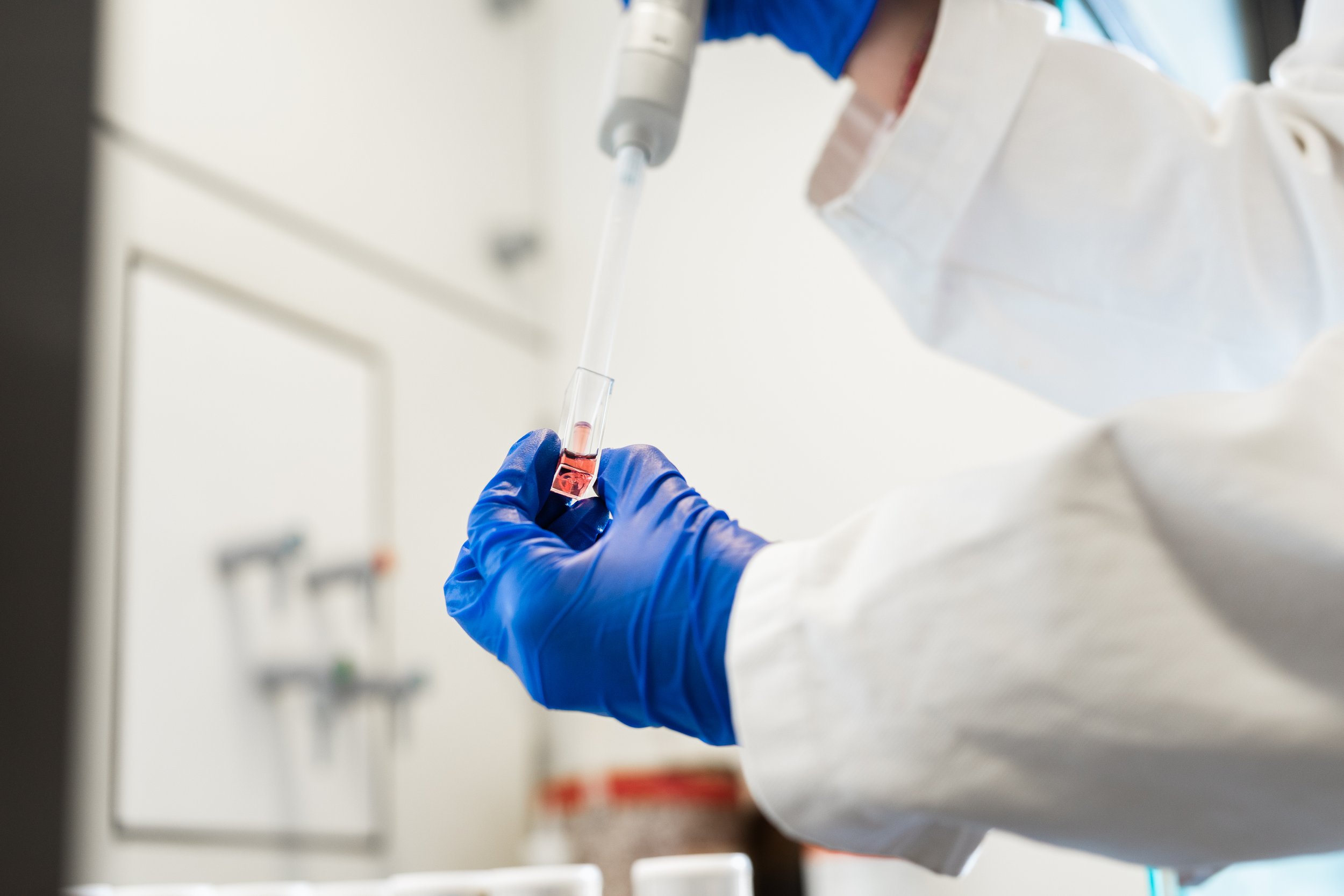 A scientist wearing blue gloves and a white lab coat is holding a test tube with a pink liquid inside in a laboratory.