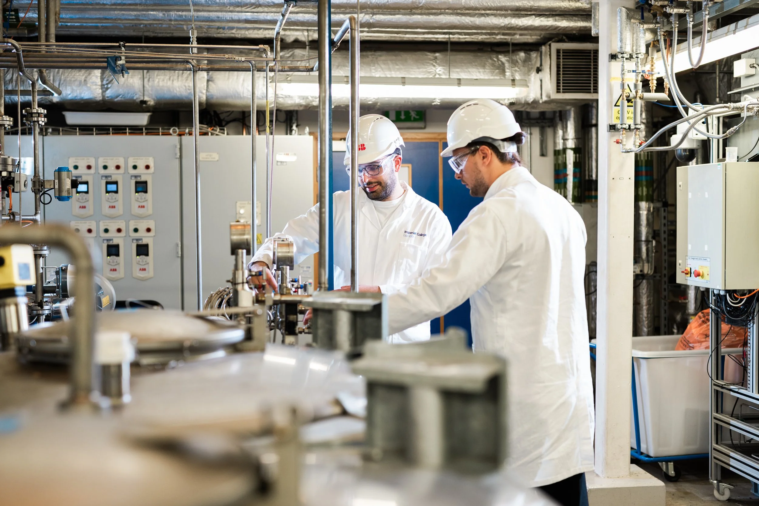 Two workers wearing white lab coats, safety helmets, and goggles working with machinery in an industrial or laboratory setting with pipes and control panels.