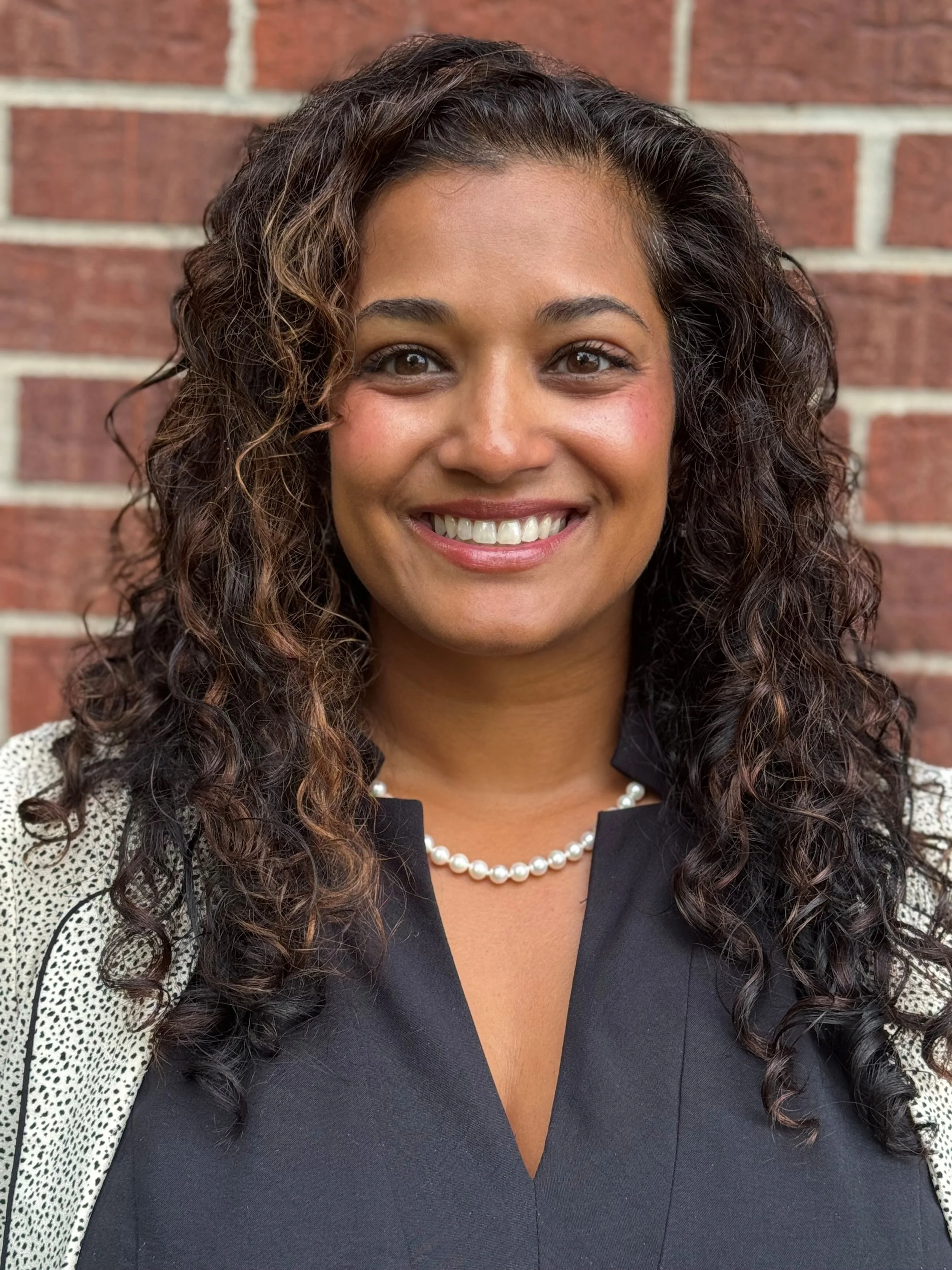 Close-up portrait of a woman with curly dark hair, smiling, standing against a brick wall, wearing a black top, a pearl necklace, and a jacket with lace details.