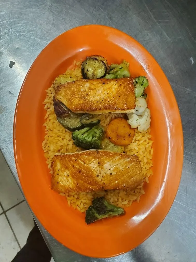 Plate of cooked yellow rice topped with two pieces of cooked fish fillets and assorted steamed vegetables including broccoli, cauliflower, zucchini, and carrots.