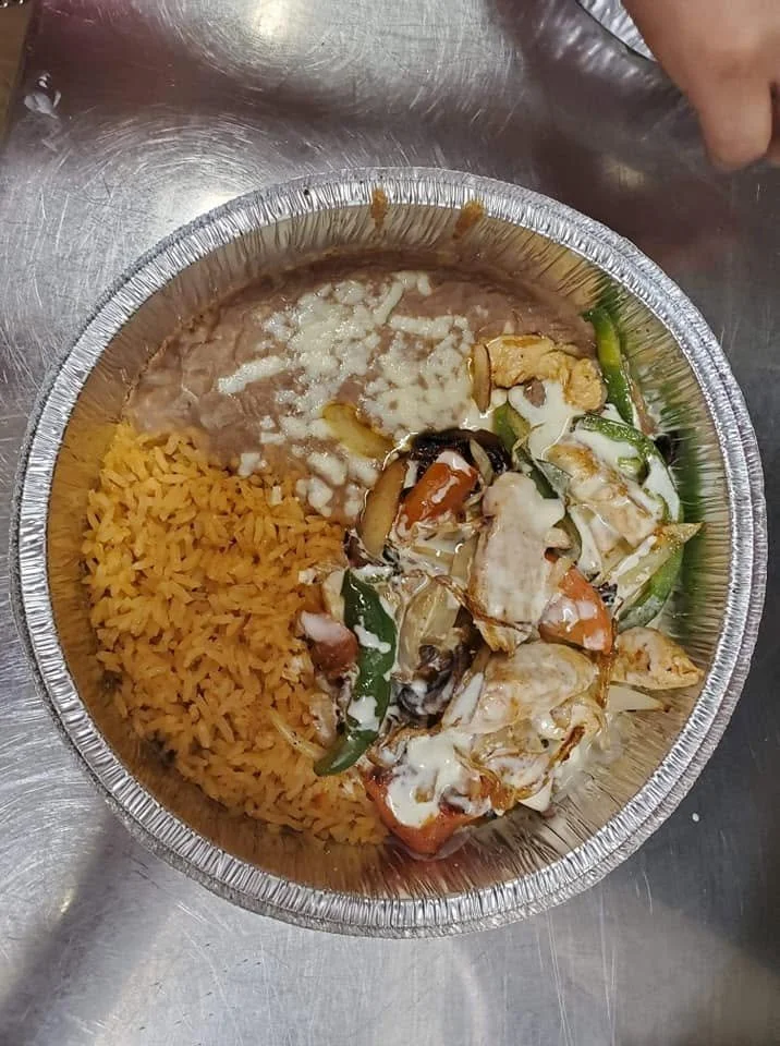 Aluminum takeout container with Mexican food including rice, refried beans, and chicken fajitas with vegetables and melted cheese, placed on a metallic surface.