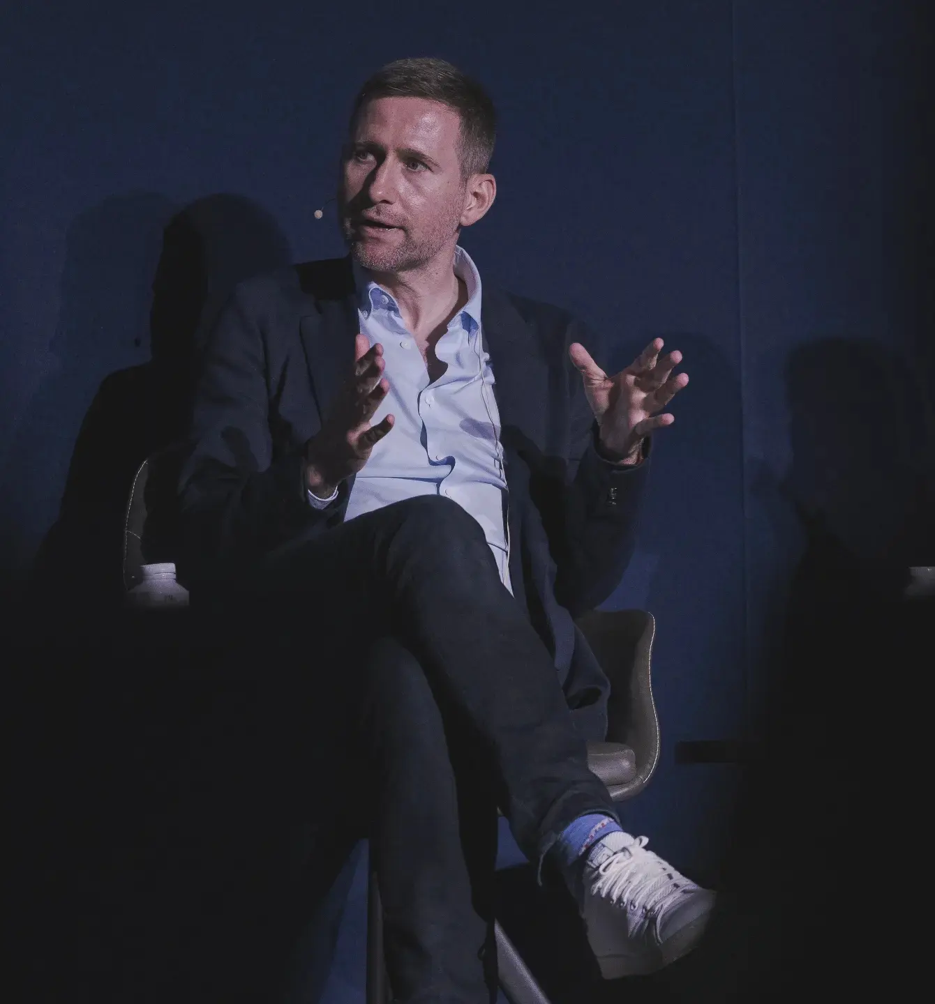 A man with light brown hair and beard, wearing a dark blazer and white shirt, seated on a chair against a dark blue background, speaking or gesturing with his hands during an interview or panel discussion.