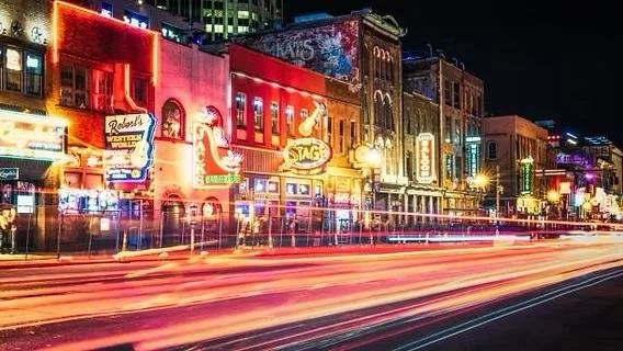 Colorful lit-up buildings and storefronts along a city street at night with light streaks from passing vehicles.