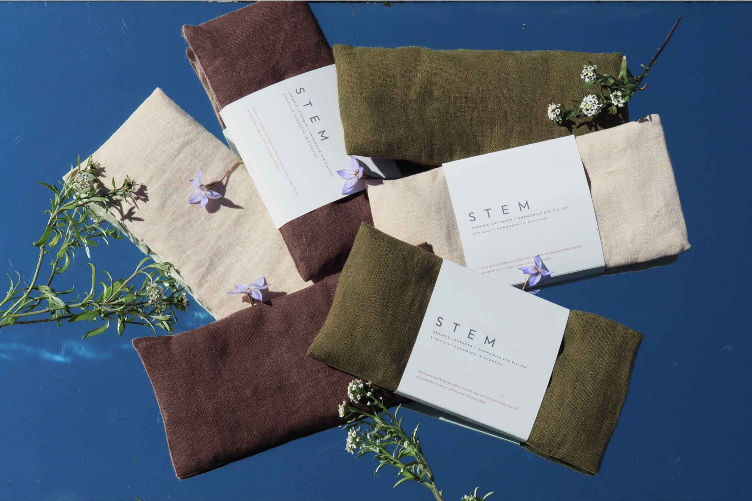 Bundles of fabric and pillowcases in earthy tones with green and purple flowers on a blue surface.