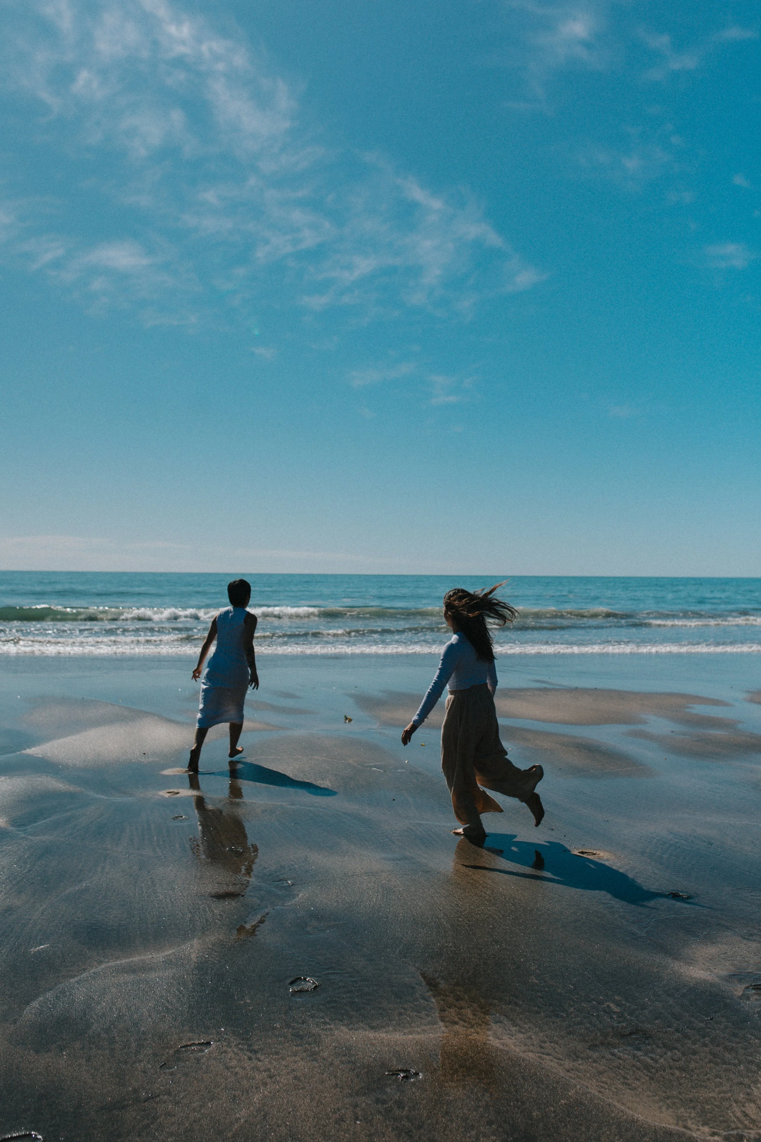 Two children playing on the sandy shoreline of a beach with waves, under a blue sky with some clouds.