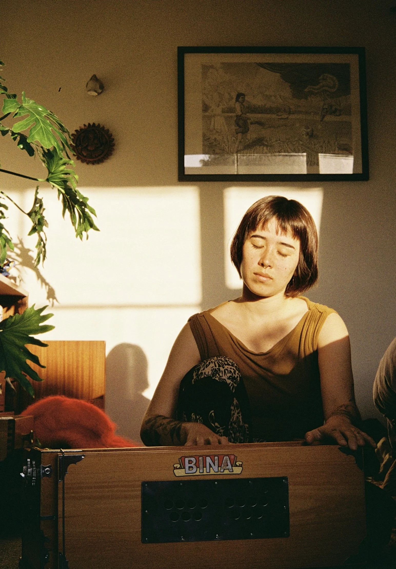 Woman with short brown hair and closed eyes playing a pinball machine named 'BINA' in a room with framed art and a plant.