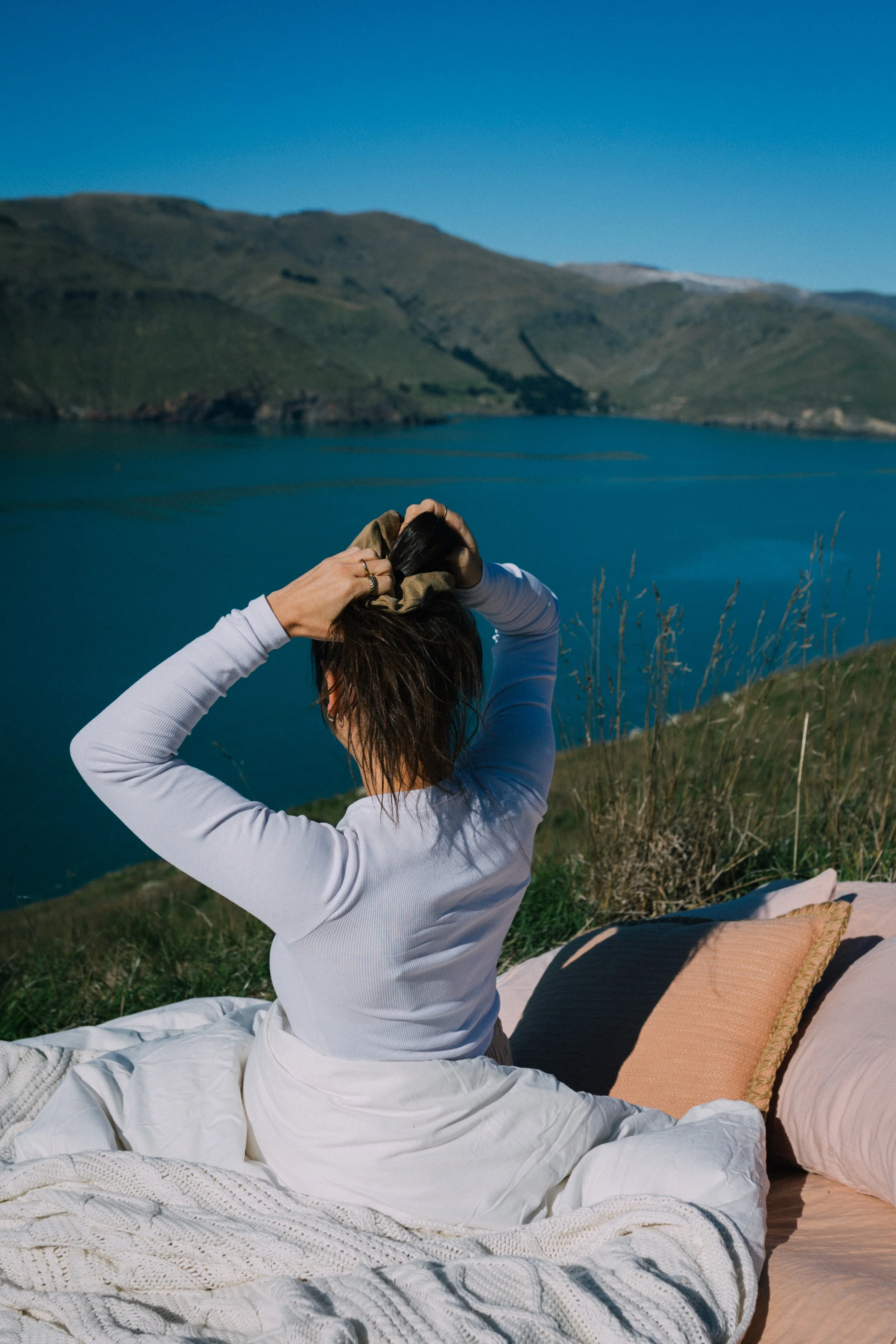Person sitting on a bed outdoors near a lake, with mountains in the background, adjusting their hair.