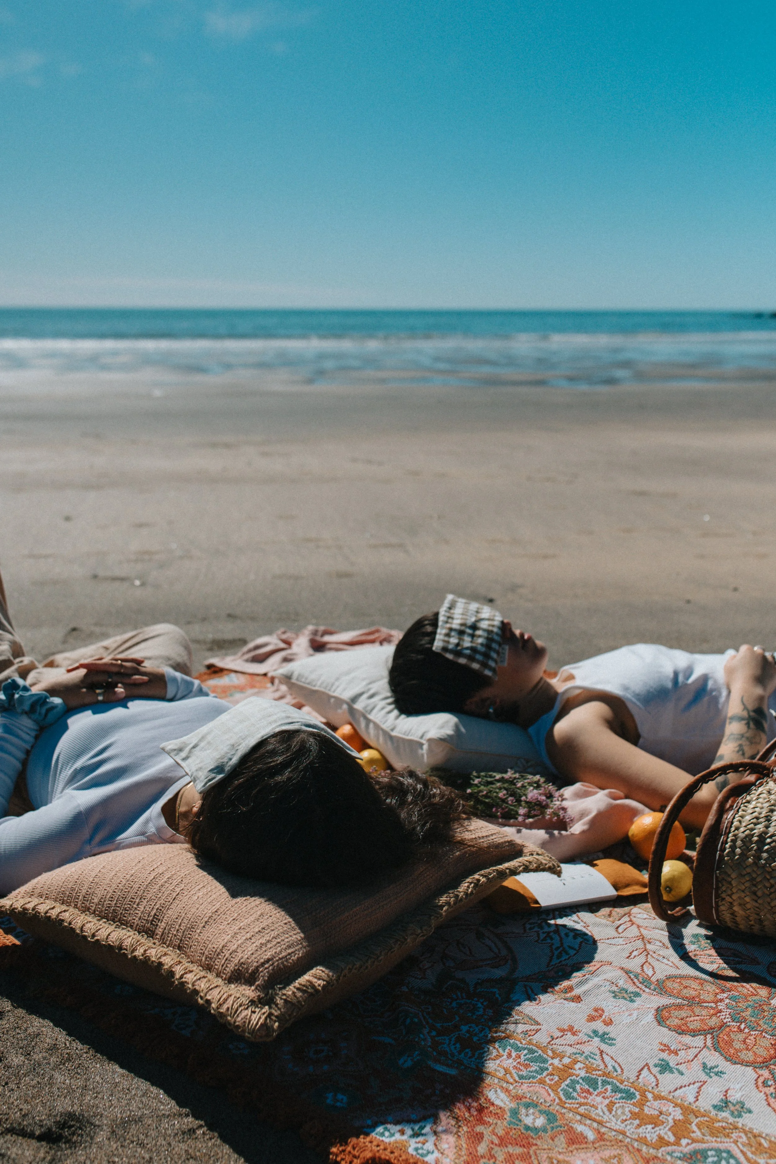 People lying on blankets and cushions on the beach, relaxing under the sun with the ocean in the background.