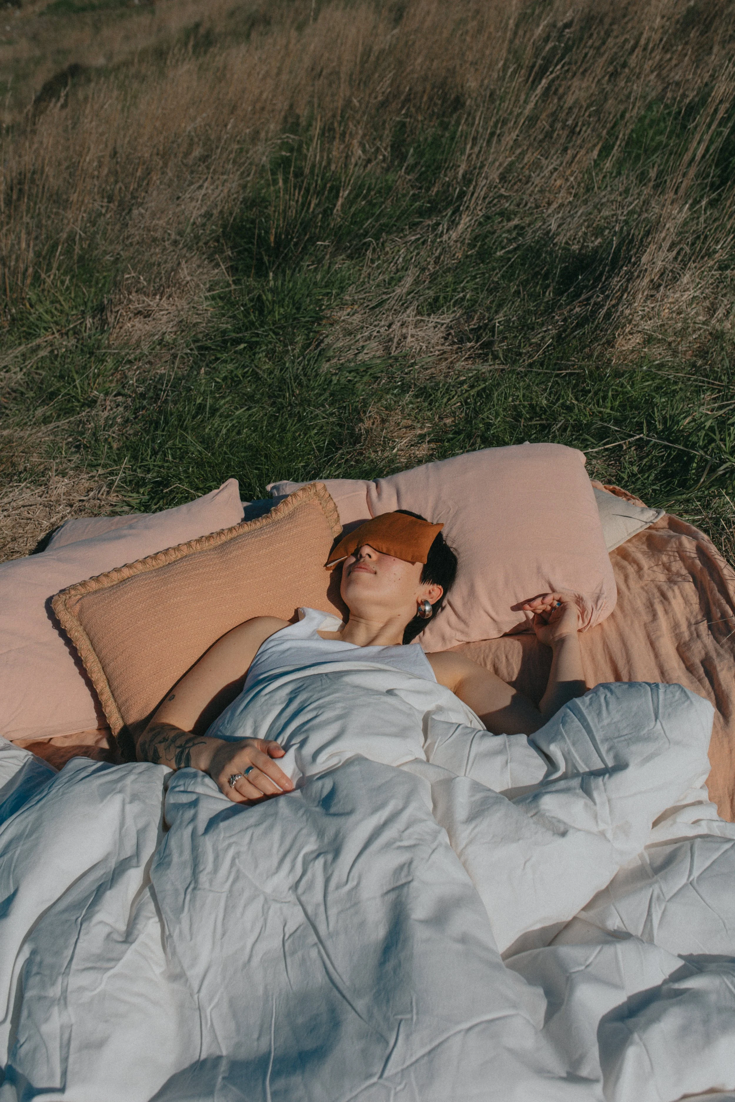 A woman lying on a bed outdoors in a grassy field, wearing a white dress and glasses, with a beige pillow and blanket.