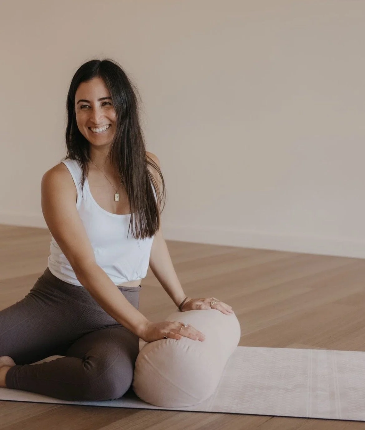 A woman practicing yoga on a mat, using a bolster pillow for support, in a room with wooden floors and neutral walls.