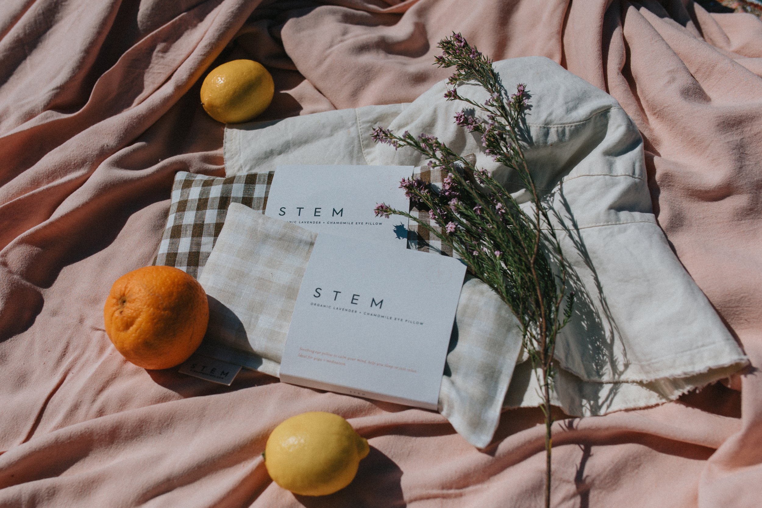 A pink fabric spread on a surface with a lemon, an orange, a white pillow with a checkered pattern, a sprig of pink flowers, and a box labeled 'STEM Organic Lavender + Chamomile Eye Pillow.'