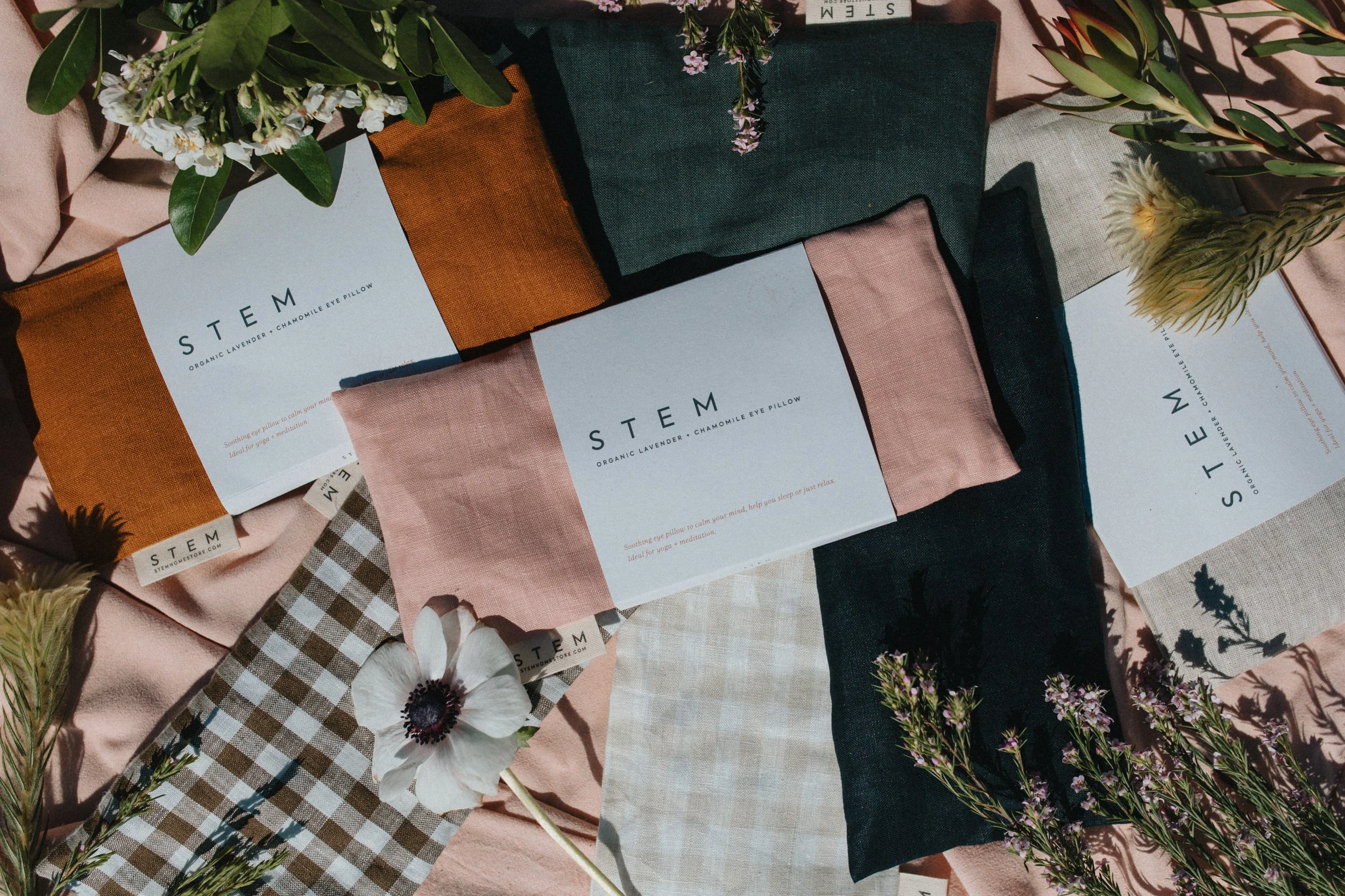 A flat lay of assorted fabric swatches and floral arrangements. The fabric swatches include pink, green, blue, orange, beige, and checkered patterns, some with tags labeled 'STEM' and describing organic lavender and chamomile eye pillows. Flowers such as white, purple, and yellow blooms are scattered among the fabrics, creating a natural and colorful display.