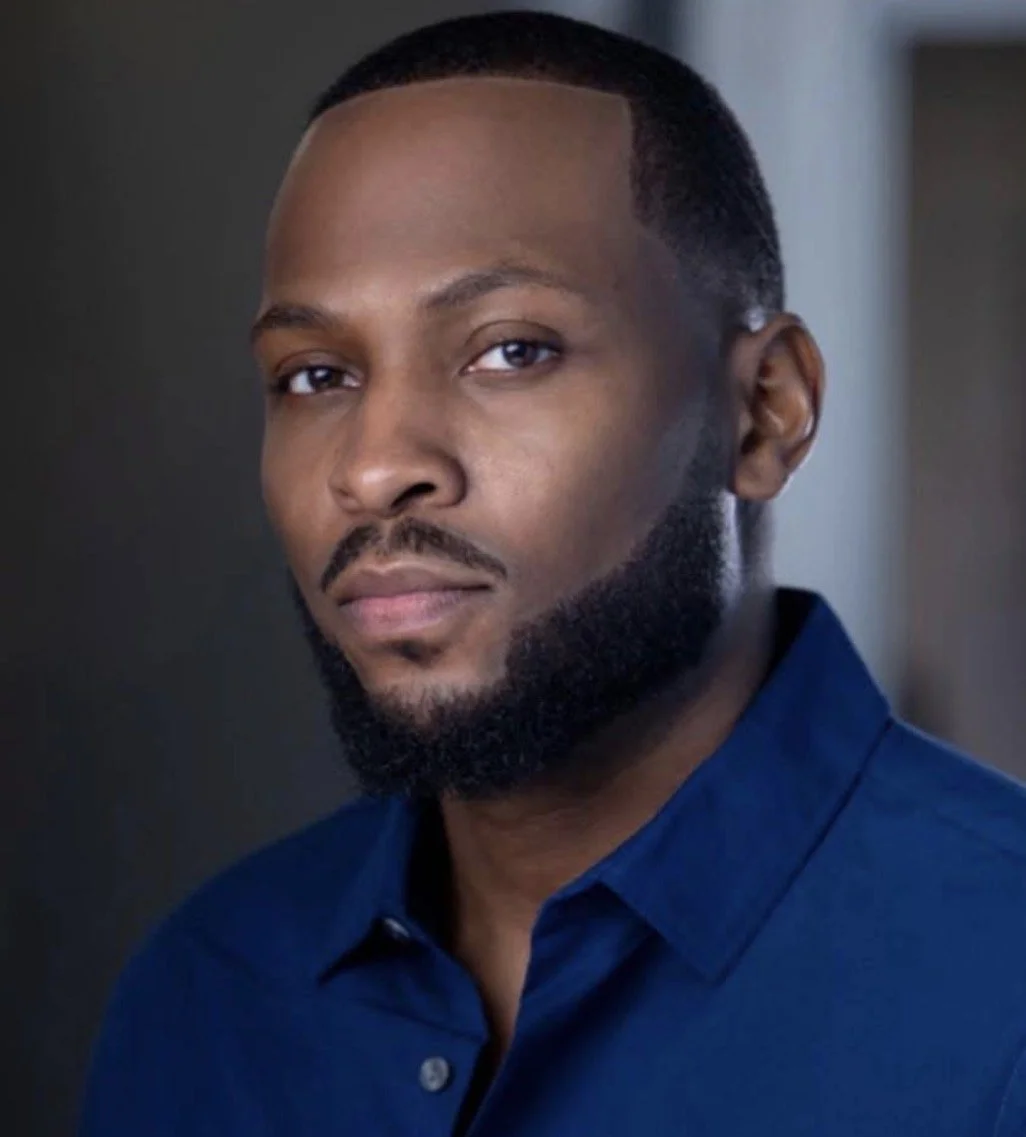 A young African American man with a beard and mustache, wearing a dark blue collared shirt, looking directly at the camera with a neutral expression.