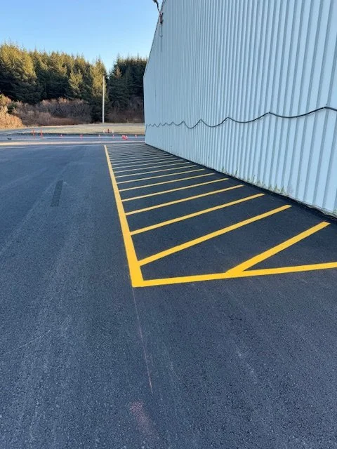 Empty parking lot with yellow diagonal lines marking a designated area, next to a large white industrial building with a corrugated metal exterior.