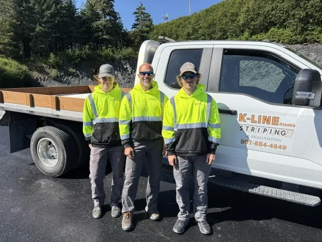 Three workers standing in front of a white company truck, wearing high-visibility yellow and black jackets, with a lush green hillside and blue sky in the background.