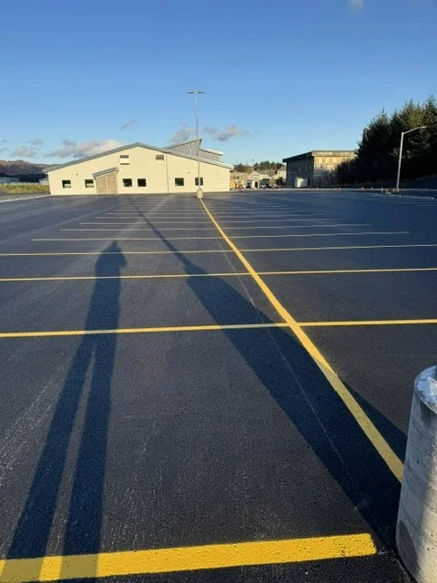 Empty parking lot with yellow lines, three buildings in the background, and long shadows cast by people taking the photo in the late afternoon or early evening.