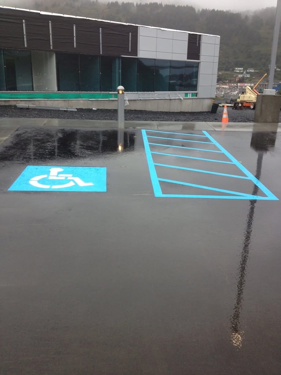 Empty parking space with bright blue lines designating a handicapped parking spot, wet pavement with puddles, and a handicapped symbol painted on the ground. In the background, a modern building, orange traffic cone, construction equipment, and a hil