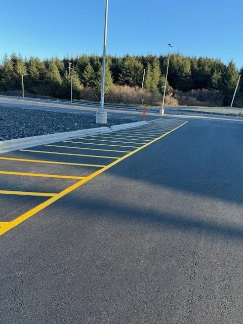 Empty parking lot with yellow parking space lines, streetlights, and a forested area in the background under a clear blue sky. Base Kodiak Line striping.