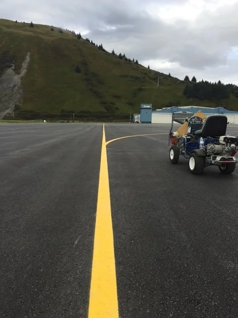 Empty airport tarmac with a yellow centerline, a remote control car, and a hillside with trees and a cloudy sky in the background.
