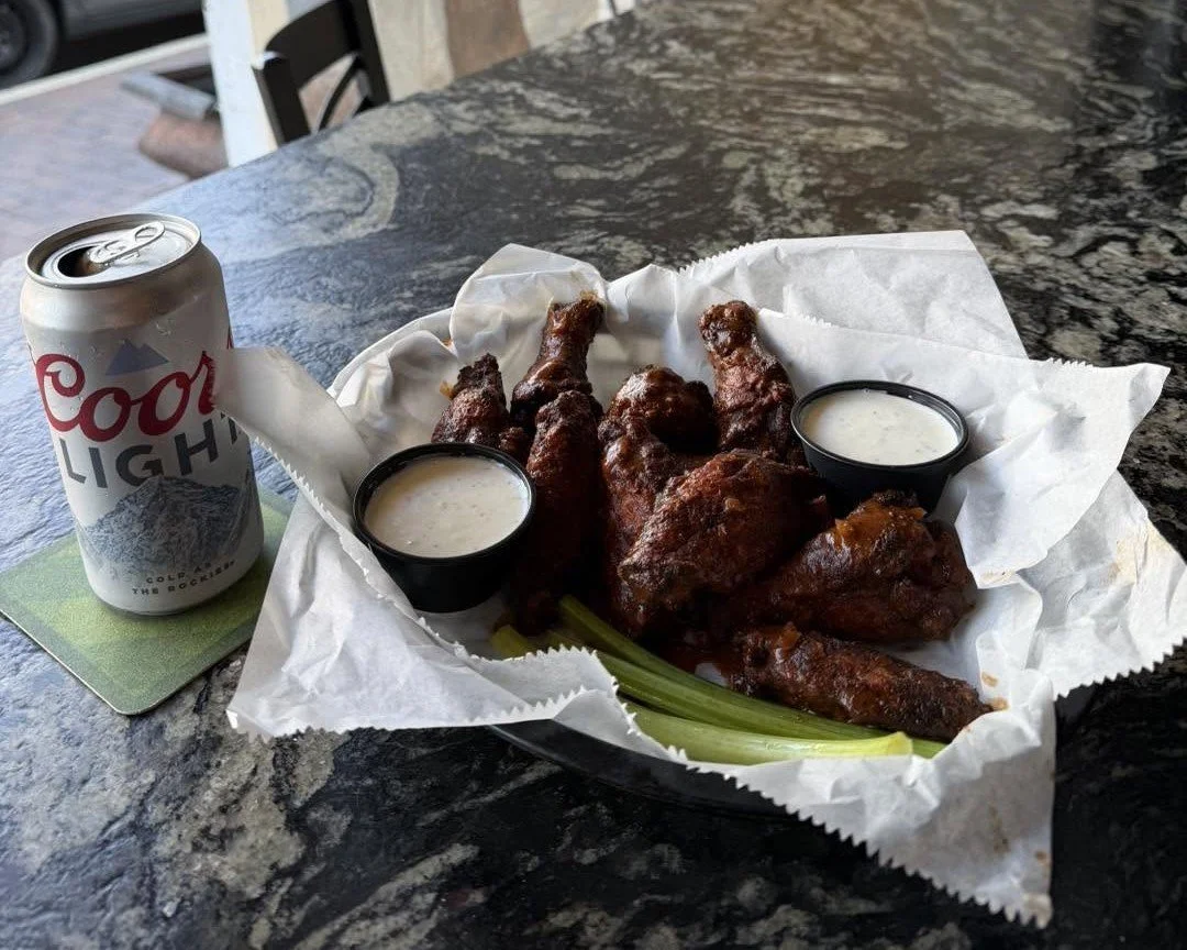 A tray of buffalo chicken wings with two cups of ranch dipping sauce and celery sticks, accompanied by a can of Coors Light beer on a marble table.
