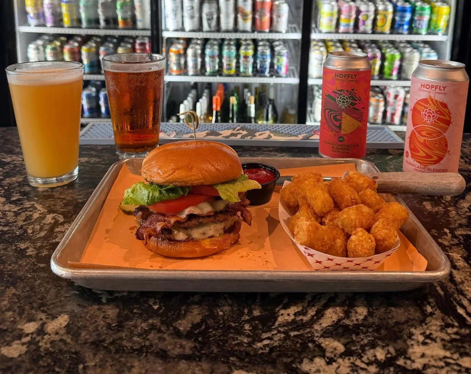 A tray with a cheeseburger, a side of tater tots, a small container of ketchup, and two beverages, in front of a refrigerator with canned drinks.
