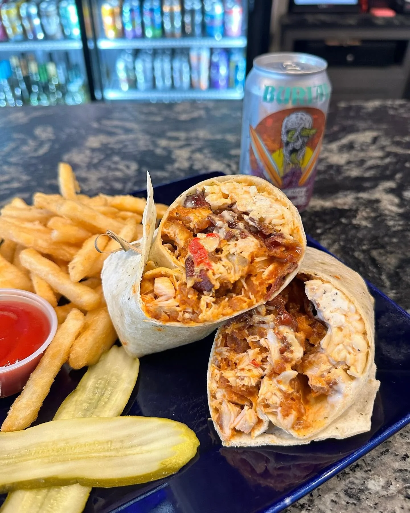 Close-up of a burrito cut in half, showing fillings of chicken, beans, cheese, and vegetables, served with French fries, pickle slices, and ketchup, with a can of soda in the background.