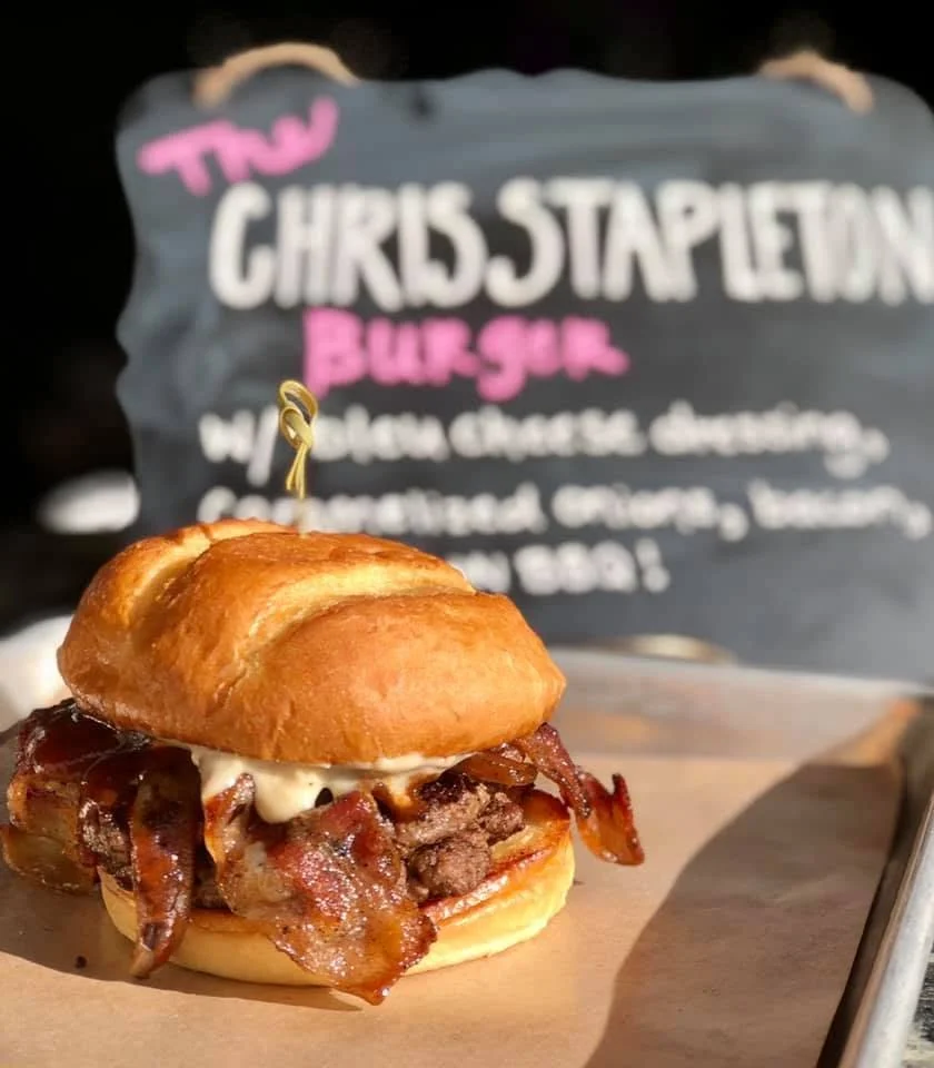 Bacon cheeseburger slider with bun, bacon, cheese, and beef patty on a small paper-lined tray.