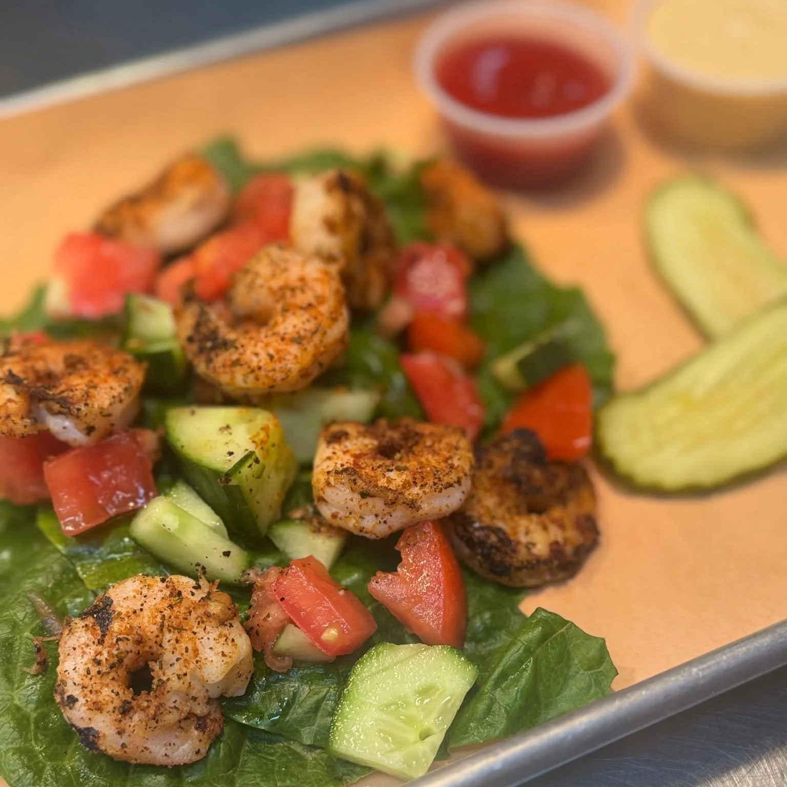 Close-up of grilled shrimp, chopped tomatoes, cucumbers, lettuce, and sliced cucumbers on a plate with a small container of red dipping sauce in the background.