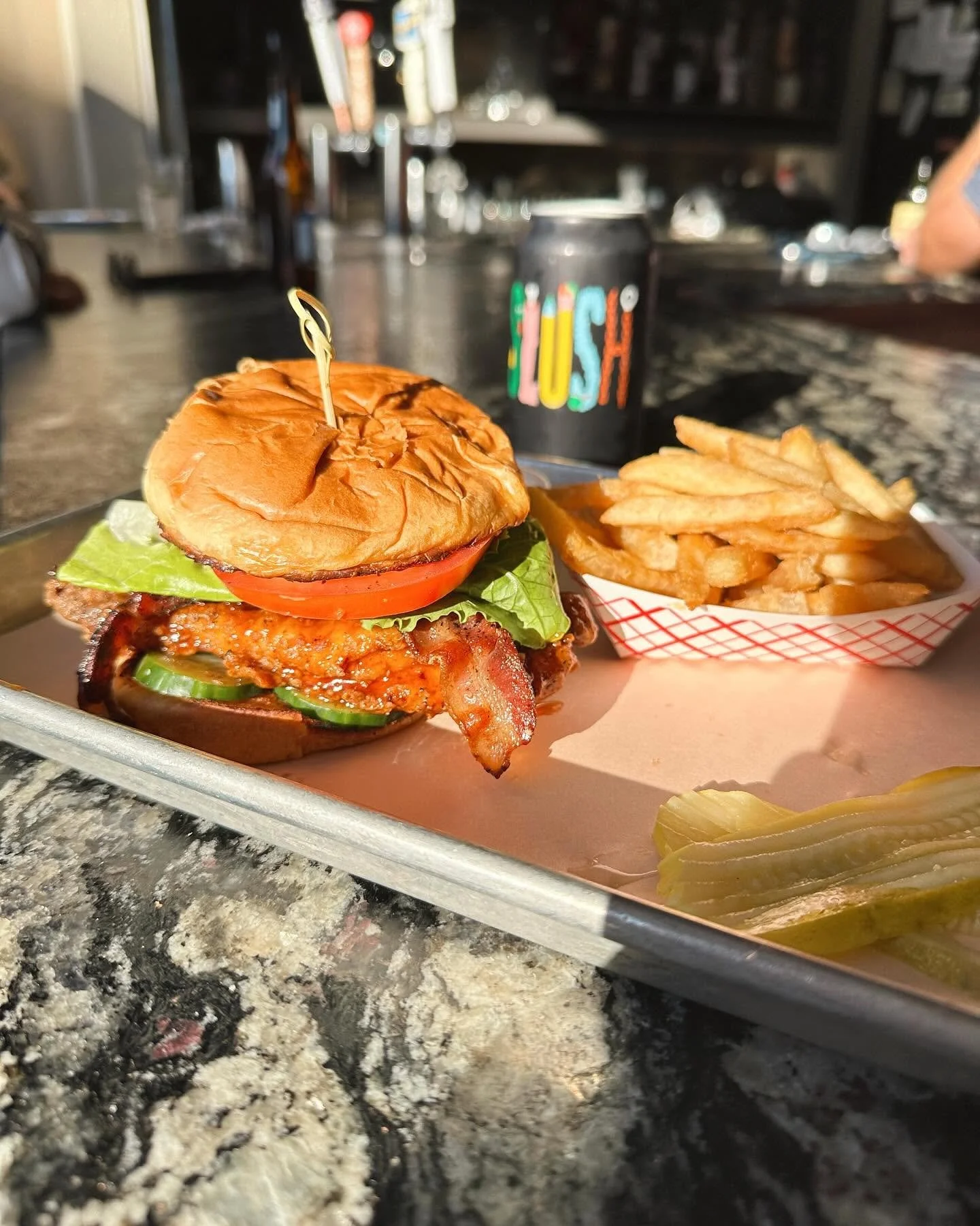 A burger with lettuce, tomato, bacon, and a sandwich bun on a tray, along with a side of French fries and pickle slices, and a dark soda can in the background.