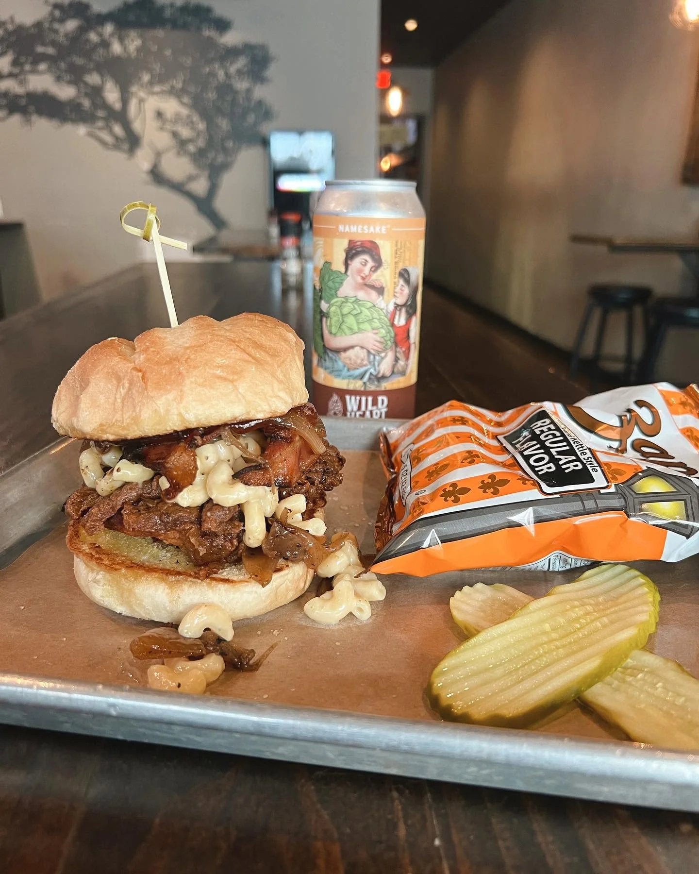 A tray of a barbecue sandwich with toppings, pickles, a bag of potato chips, sliced pickles, a canned beverage, and pickles on a wooden table inside a restaurant with a wall mural of a tree in the background.