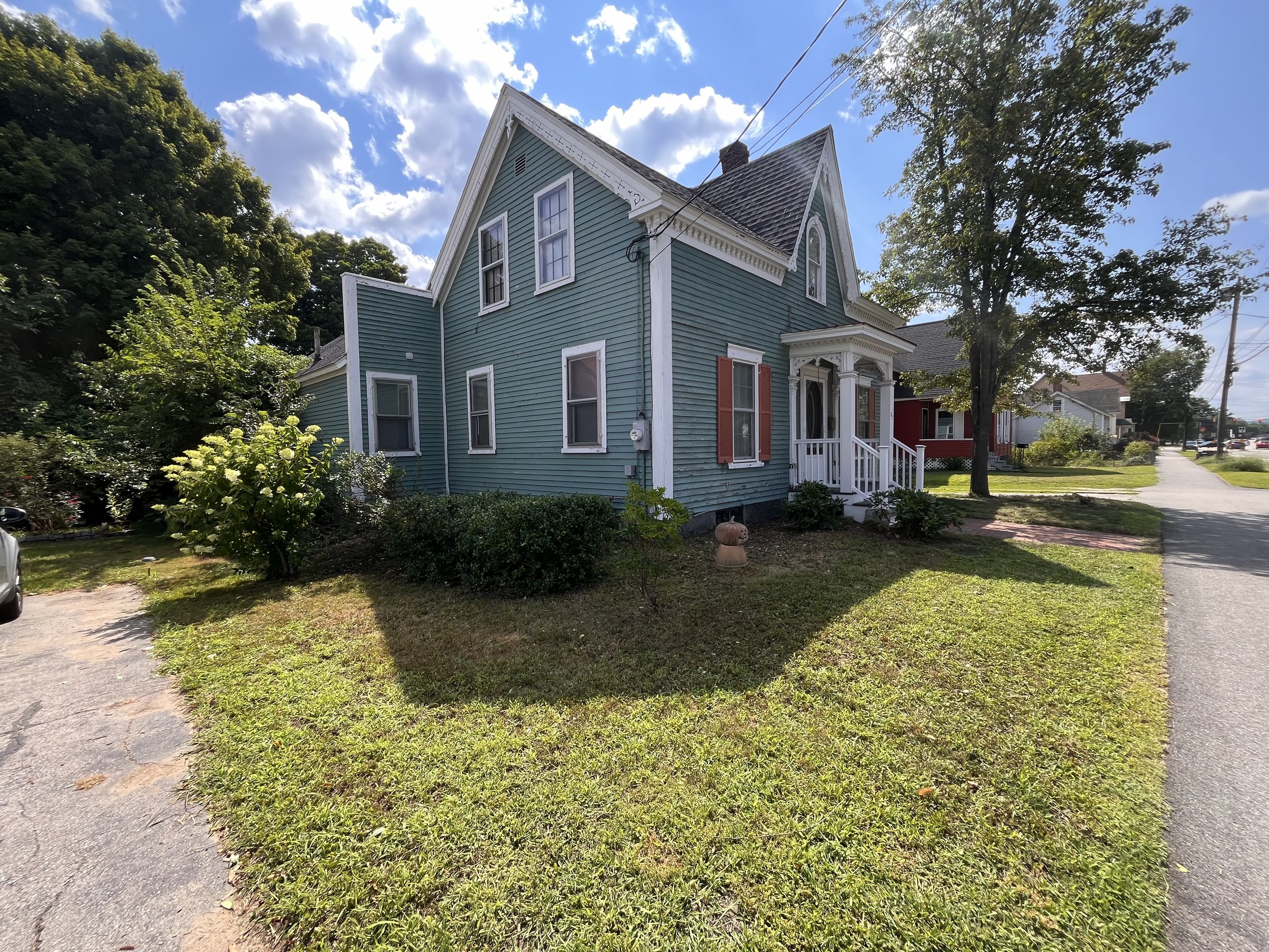 A blue house with white trim and orange shutters on a sunny day with a partly cloudy sky. There are trees and a sidewalk nearby.