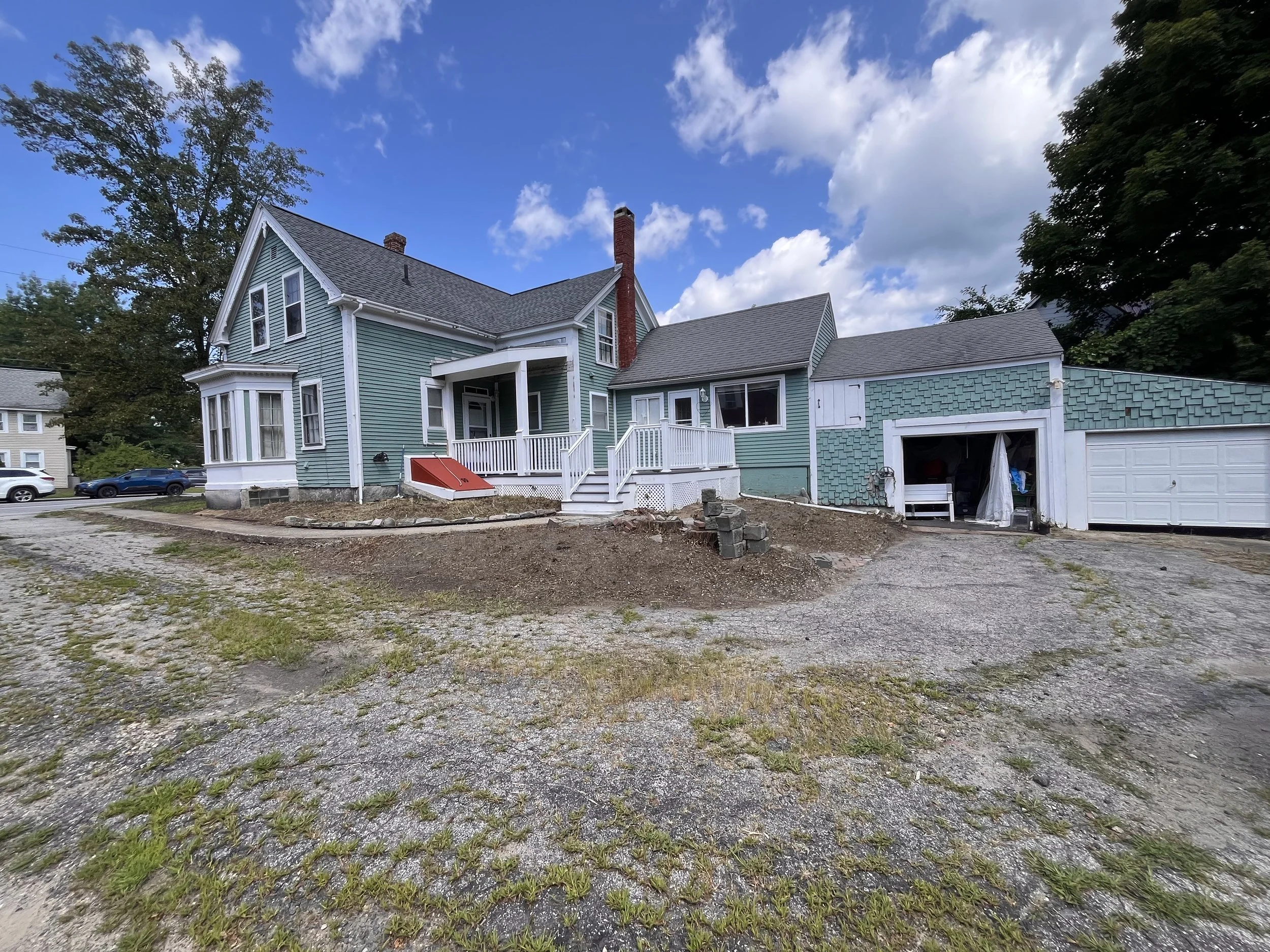 A house with blue siding, a porch, and a garage under construction, surrounded by a gravel driveway and trees, with a partly cloudy sky overhead.