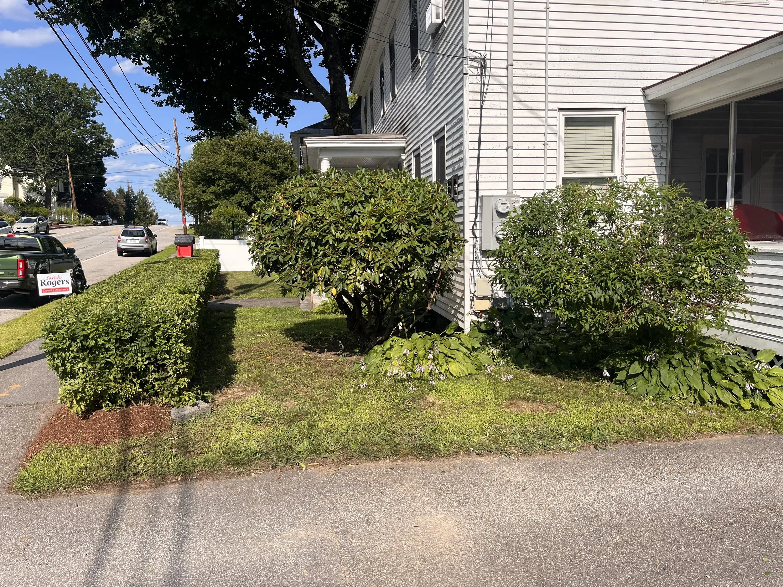 A residential street scene with parked cars, green bushes, trees, sidewalk, and a white house with siding. The sky is blue with some clouds.